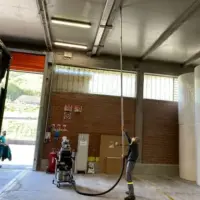 Operator uses a Depureco ECOBULL industrial vacuum with a long-reach hose to remove dust from overhead facility surfaces.