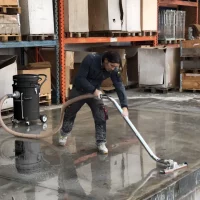Man using an industrial liquid vacuum with an S-pipe and liquid floor brush to clean a large spill on a warehouse concrete floor.